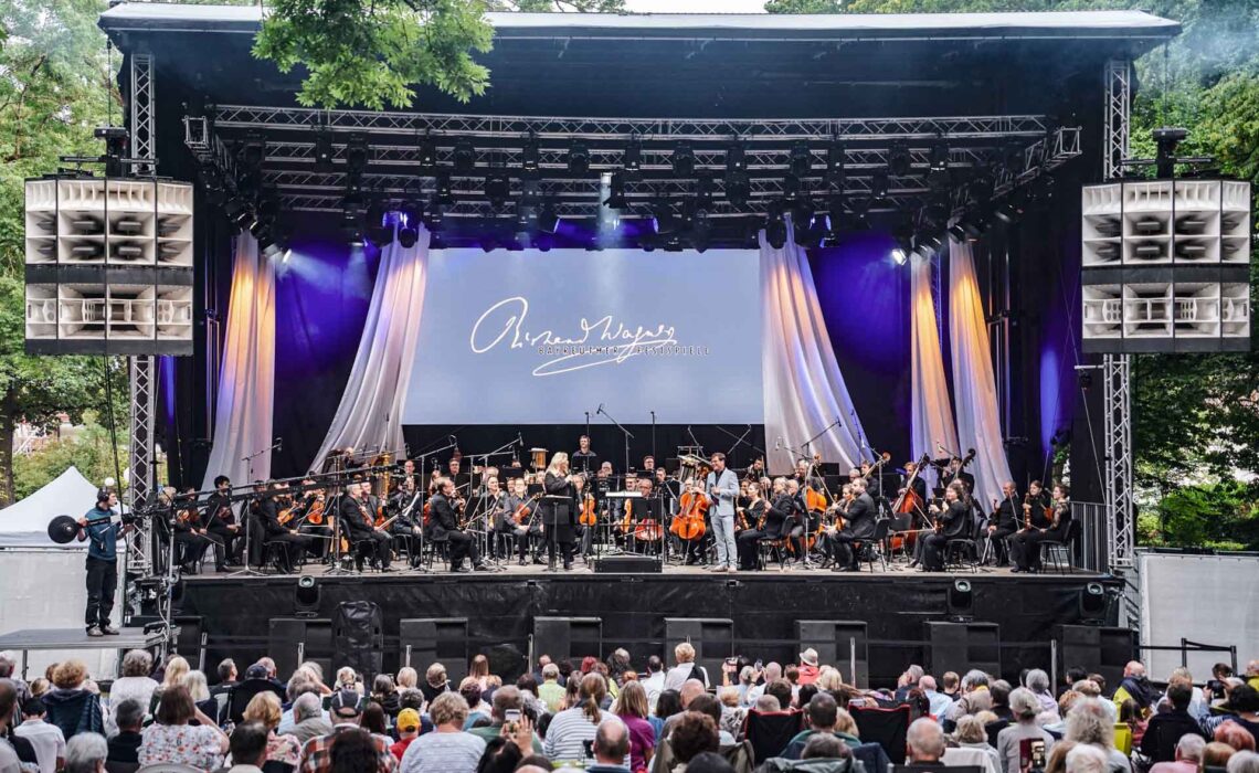 Mit Geldermann bei den Bayreuther Festspielen 2025: Festspiel-Open-Air im Festspielpark. Auf der Bühne ist das Festspielorchester zu sehen, davor Moderator Axel Brüggemann im Gespräch mit der Festspielchefin der Bayreuther Festspiele, Katharina Wagner. Im Vordergrund ist das Publikum im voll besetzten Festspielpark zu sehen