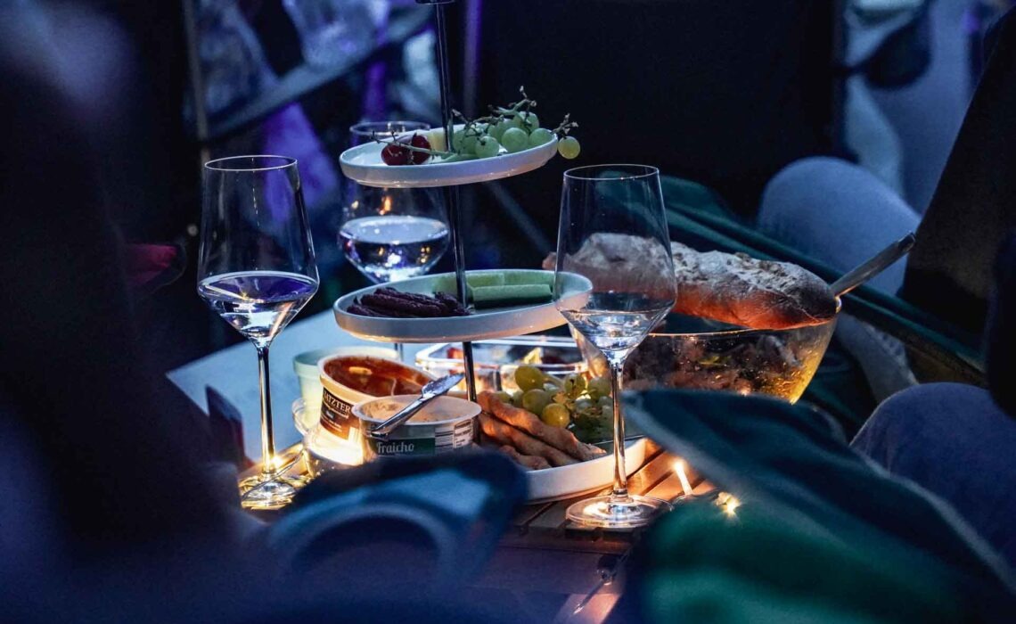 Mit Geldermann bei den Bayreuther Festspielen 2025: Festspiel-Open-Air im Festspielpark. Blick ins Publikum auf eine Picknickdecke mit Lichterkette, Etagere, Käse, Baguette, Weintrauben und Wein bei Nacht