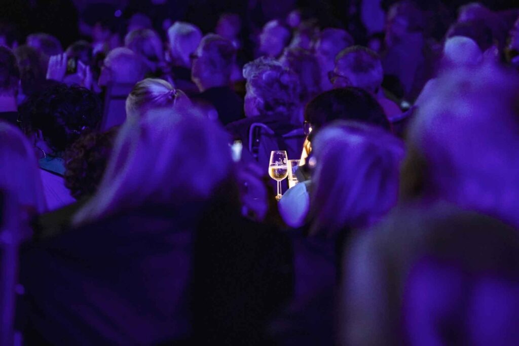 Mit Geldermann bei den Bayreuther Festspielen 2025: Festspiel-Open-Air im Festspielpark. Blick ins Publikum bei Nacht und mit blauer Beleuchtung auf einen Tisch mit einer kleinen Tischleuchte und einem Weinglas