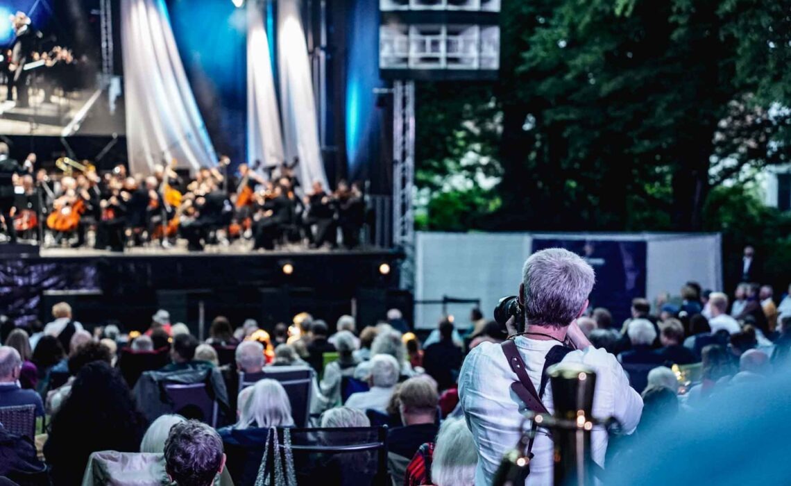 Mit Geldermann bei den Bayreuther Festspielen 2025: Festspiel-Open-Air im Festspielpark. Blick über die Schulter eines Pressefotografen über das Publikum hinweg auf die Bühne und das Festspielorchester