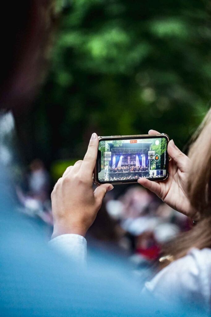 Mit Geldermann bei den Bayreuther Festspielen 2025: Blick durch das Smartphone einer Besucherin auf die Bühne und das Festspielorchester beim Festspiel-Open-Air im Festspielpark