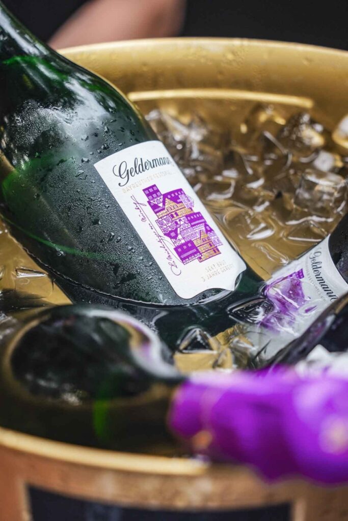 Mit Geldermann bei den Bayreuther Festspielen 2025: Die Geldermann Festspiel-Cuvée mit dem Motiv zu die Meistersinger von Nürnberg mit typischen Fachwerkhäusern in Lila und Gold in einem Sektkühler mit Eiswürfeln in der Geldermann-Lounge beim Festspiel-Open-Air im Festspielpark