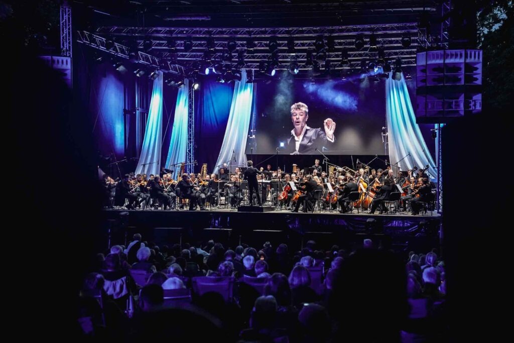 Mit Geldermann bei den Bayreuther Festspielen 2025: Festspiel-Open-Air im Festspielpark. Blick auf die Bühle und das Festspielorchester bei Nacht mit spektakulärer Beleuchtung in Blau. Auf einem großen Screen im Hintergrund ist der spanische Dirigent Pablo Heras-Casado in Großaufnahme zu sehen