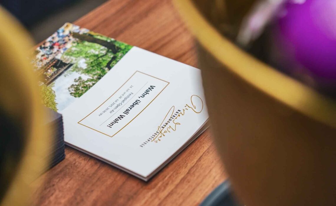 Mit Geldermann bei den Bayreuther Festspielen 2025: Festspiel-Open-Air im Festspielpark. Sektbar in der Geldermann-Lounge. Auf der Theke liegt ein Programmheft in Weiß mit dem Konzerttitel Wahn, überall Wahn, davor ist ein Sektkühler in Gold zu sehen