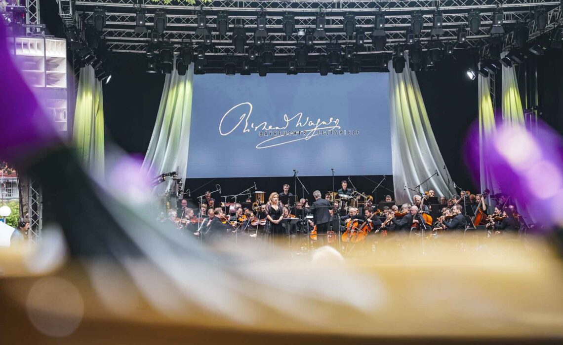 Mit Geldermann bei den Bayreuther Festspielen 2025: Festspiel-Open-Air im Festspielpark. Blick von der Geldermann-Lounge über einen Sektkühler mit Sektflaschen auf die Bühne. Aufführung des Festspiel-Orchesters und Sopranistin Gabriela Scherer. Im Hintergrund ein blauer Screen mit dem Schriftzug Richard Wagners in Gold