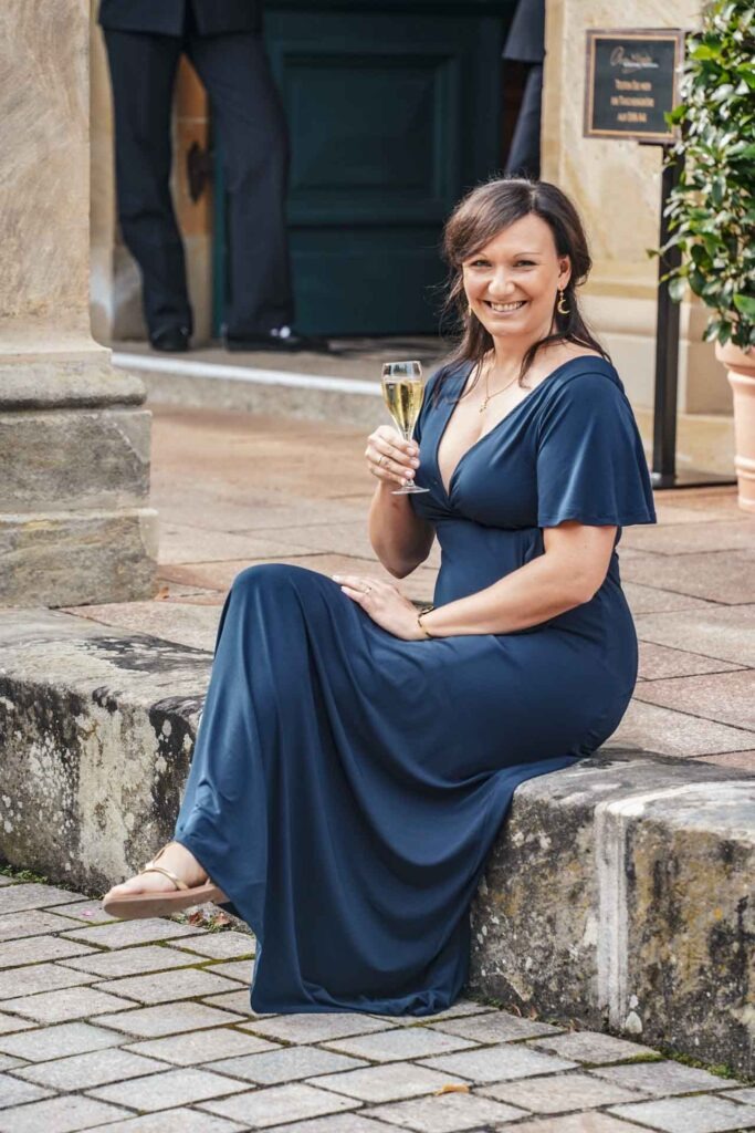 Mit Geldermann bei den Bayreuther Festspielen 2025: Content Creatorin Alexandra Baier von Tables & Fables sitzt auf einer Mauer vor dem Eingang zur Mittelloge des Bayreuther Festspielhauses. Sie trägt ein dunkelblaues Abendkleid, hält ein Glas der Geldermann Bayreuther Festspiel-Cuvée in der Hand und lächelt in die Kamera