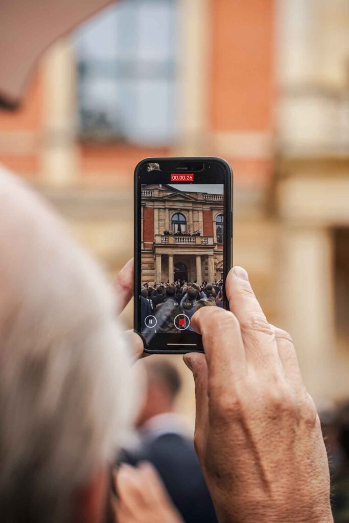 Mit Geldermann bei den Bayreuther Festspielen 2025: Blick über die Schulter einer ältern Festspielbesucherin, die ein Video der Pausenmusiker auf dem Balkon des Festspielhauses mit ihrem Smartphone aufzeichnet