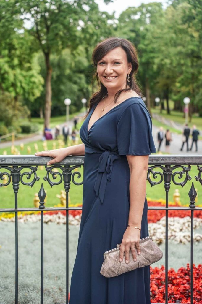 Mit Geldermann bei den Bayreuther Festspielen 2025: Content Creatorin Alexandra Baier im dunkelblauen Abendkleid und Clutch in Gold auf dem Grünen Hügel in Bayreuth, im Hintergrund der Festspielpark mit Blumenbeeten und den Wagner-Skulpturen von Ottmar Hörl