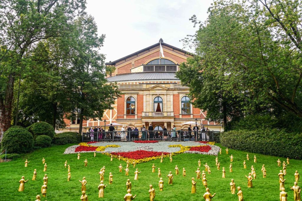 Mit Geldermann bei den Bayreuther Festspielen 2025: Blick vom Festspielpark auf das Bayreuther Festspielhaus. Davor sieht mein eine Skulptureninstallation des Künstlers Ottmar Hörl mit kleinen Richard-Wagner-Figuren (Modell Die Verneigung) sowie Figuren von König Ludwig II. (Modell Der Mäzen) in Gold vor dem großen Blumenbeet