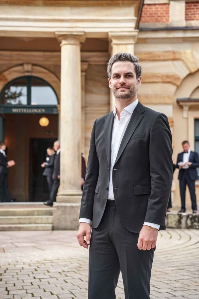 Mit Geldermann bei den Bayreuther Festspielen 2025: Florian Baier von Tables & Fables im schwarzen Anzug vor dem Eingang zur Mittelloge des Bayreuther Festspielhauses