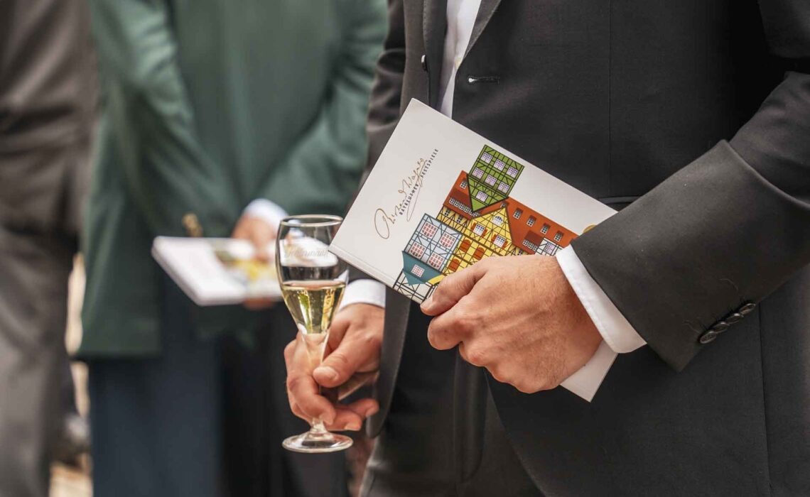 Mit Geldermann bei den Bayreuther Festspielen 2025: Nahaufnahme von Florian Baier von Tables & Fables im Anzug auf dem Vorplatz des Bayreuther Festspielhauses. Er trägt einen schwarzen Anzug und hält in der einen Hand das Programmheft zu Die Meistersinger von Nürnberg, in der anderen Hand ein Glas Sekt mit Geldermann Bayreuther Festspiel-Cuvée