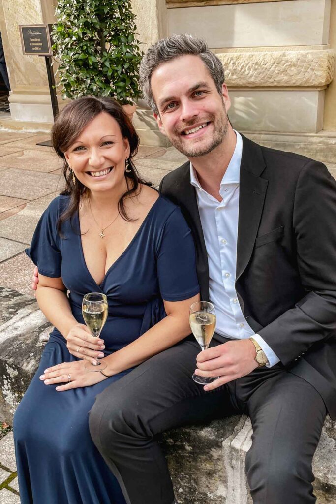 Mit Geldermann bei den Bayreuther Festspielen 2025: Content Creatorin Alexandra Baier und Florian Baier von Tables & Fables sitzen vor dem Festspielhaus auf einer Steinmauer. Alexandra trägt ein dunkelblaues Abendkleid, Florian einen schwarzen Anzug. Beide lächeln in die Kamera und halten ein Glas Geldermann Sekt mit Bayreuther Festspiel-Cuvée in der Hand