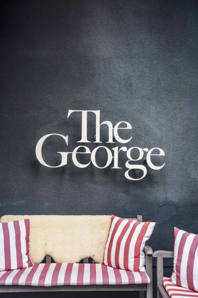 Hoteltipps Hamburg: Schriftzug in Weißen Lettern auf der grauen Außenfassade des The George Hotel, darunter bequeme Lounge Sessel mit rot-weißen Streifen
