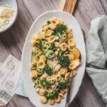 Orecchiette mit Brokkoli und weißen Bohnen im Flatlay-Stil auf einer weißen Servierplatte auf Holzbrett und Holztisch. Gehobelter Parmesan, Wasserglas, hellgrüne Serviette und Vintage-Postkarten schaffen eine mediterrane Tischszene inspiriert von der Küche Apuliens